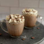 Two clear mugs of freshly-made hot cocoa topped with whipped cream and chocolate shavings.