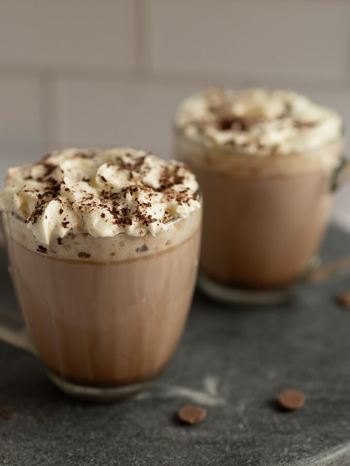 Two clear mugs of freshly-made hot cocoa topped with whipped cream and chocolate shavings.