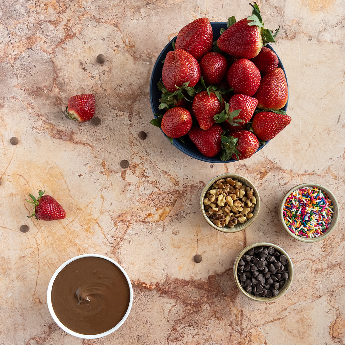 A big bowl of fresh strawberries and tempered milk chocolate, alongside three bowls of various toppings.