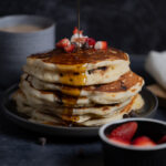 A stack of chocolate chip pancakes with a fresh drizzle of maple syrup, served with coffee and slices strawberries.